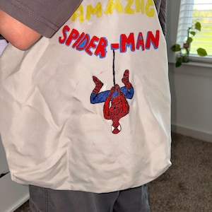 May include: Off-white tote bag featuring the words "AMAZING SPIDER-MAN" in yellow and red lettering. The bag has an embroidered image of Spider-Man hanging upside down. The bag is made of a soft, durable fabric.