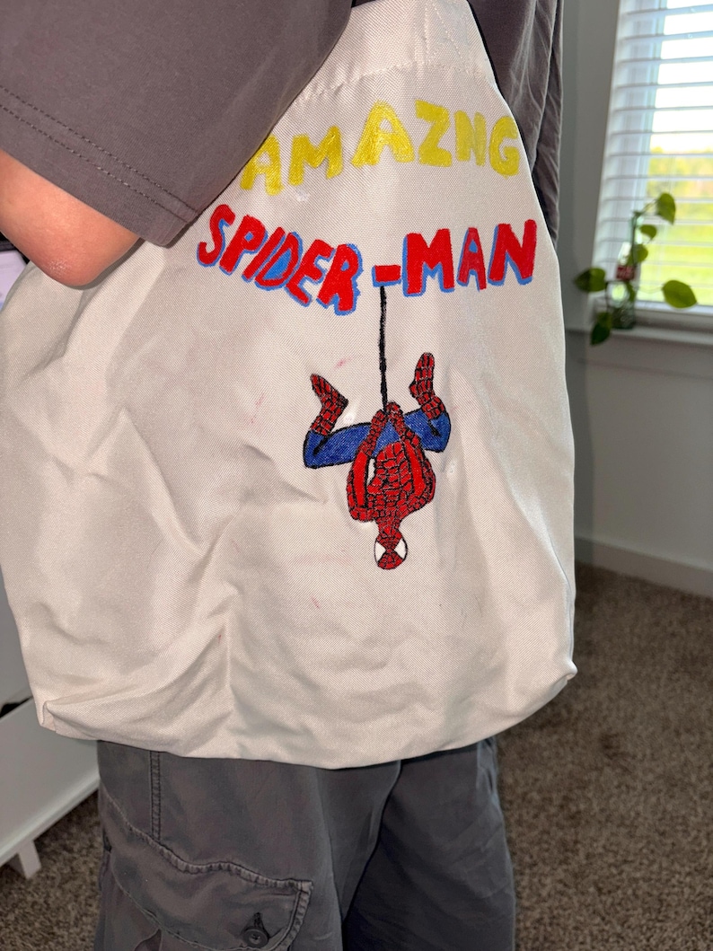 May include: Off-white tote bag featuring the words "AMAZING SPIDER-MAN" in yellow and red lettering. The bag has an embroidered image of Spider-Man hanging upside down. The bag is made of a soft, durable fabric.