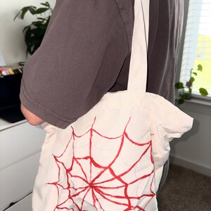 May include: A white tote bag with a red spiderweb design. The bag has a rectangular shape and two white straps. The spiderweb design is centered on the front of the bag.
