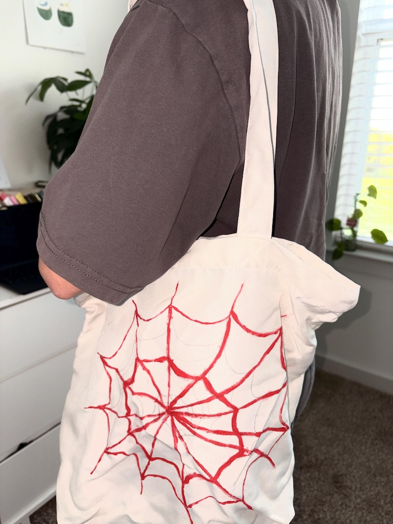 May include: A white tote bag with a red spiderweb design. The bag has a rectangular shape and two white straps. The spiderweb design is centered on the front of the bag.