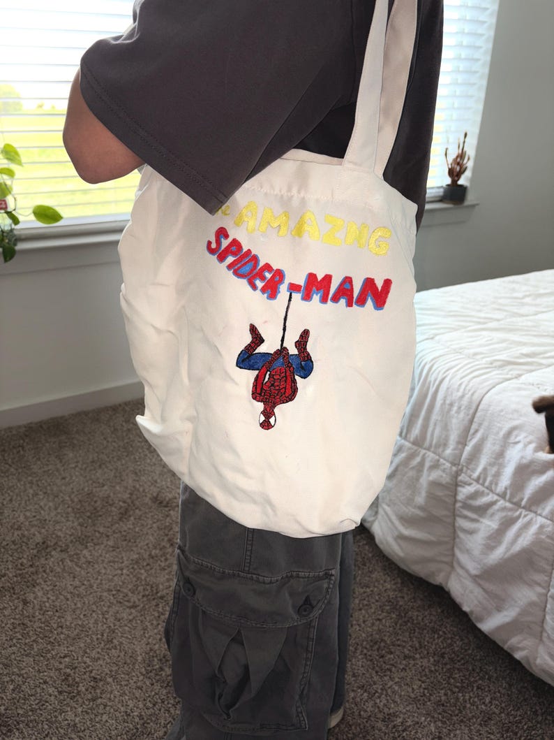 May include: White tote bag with cream-colored straps featuring the text "AMAZING SPIDER-MAN" in yellow and red embroidery. The bag has an embroidered image of Spider-Man hanging upside down.