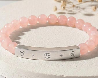 May include: A pink rose quartz bracelet with a silver-coloured bar engraved with zodiac symbols. The bracelet is presented on a white surface. Text at the bottom reads: SELECT FROM 7 STONE TYPES, ENGRAVE YOUR SUN, MOON, RISING SIGNS, STAINLESS STEEL OR 18K GOLD PLATED BAR.