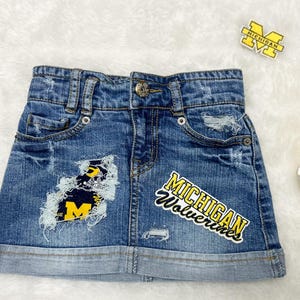 May include: Blue denim skirt with distressed details and patches featuring the University of Michigan logo and "Michigan Wolverines" text. Includes a foam finger, a drink with a straw, and the word "Michigan".