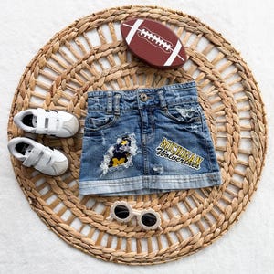 May include: A blue denim skirt with distressed detailing, a Michigan Wolverines patch, and a frayed fabric accent. Accompanied by a brown football, a pair of white sneakers with hook-and-loop straps, and round cream-colored sunglasses.