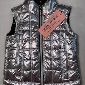 May include: Reversible silver puffer vest with a quilted design and a black interior. The vest has a full zipper closure and a tag that reads "REVERSIBLE URBAN REPUBLIC". The vest is part of the girls collection.