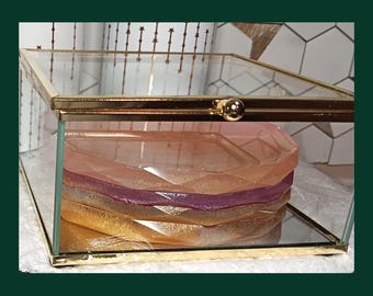 Puede incluir: Una caja de exhibición de vidrio transparente y metal dorado con una pila de piezas de resina coloridas y de forma geométrica en su interior. Las piezas de resina son en tonos dorados, rosas y morados. La caja tiene una tapa con bisagras y un cierre dorado.