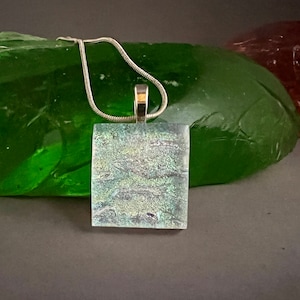 May include: A silver chain necklace with a square pendant made of iridescent glass. The pendant has a light blue and white color scheme with a subtle shimmer.