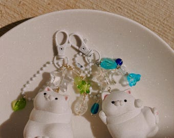 May include: Two white cat-shaped charms attached to white plastic lobster clasps. Each charm features a matte white cat figure with small pink ears and paws. The keychains are decorated with clear star-shaped beads, small green leaf beads, and blue translucent beads.