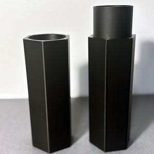 May include: Two black, hexagonal candle holders. One is a simple, open-top design, while the other features a cylindrical top section. Both are made of a matte black material and stand upright on a gray surface.