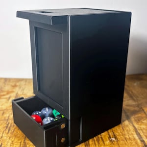 May include: A black dice tower with a pull-out tray containing various colored dice. The tower has a rectangular design with a top overhang and a slot for dice to be rolled. The tray is open, revealing the dice.