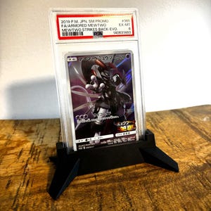 May include: A graded Pokémon trading card featuring Armored Mewtwo, displayed in a clear protective case. The card is supported by a black stand on a wooden surface. The card's details include text in Japanese and English.