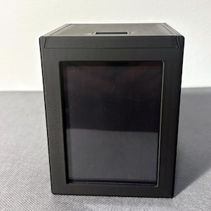Pode incluir: Uma caixa de exibição preta em forma de cubo com uma janela de visualização retangular. A caixa tem um acabamento fosco e uma pequena ranhura na parte superior. A janela é escura e a caixa está sobre uma superfície texturizada.