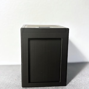 May include: A black, square box with a recessed panel on the front. The box has a matte finish and a small rectangular opening on the top. The object is sitting on a gray surface.