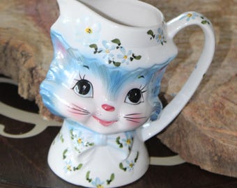 May include: A white ceramic creamer shaped like a cat's head, with blue fur and floral accents. The cat has large black eyes, pink cheeks, and a bow tie. The handle and rim are also decorated with flowers. A charming addition to any kitchen.