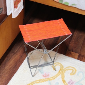 May include: An orange folding stool with a metal frame and chain supports. The seat is made of orange fabric with white stitching. A white towel with a camper graphic hangs in the background.