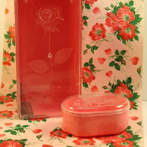 May include: Two pink plastic boxes with a rose design. The larger box is rectangular and has a clear lid. The smaller box is oval and has a lid with a rose design. Both boxes are sitting on a pink and green floral patterned paper.