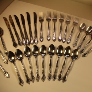 May include: A set of silverware with a silver finish, including forks, knives, and spoons. The silverware is arranged in a fan shape on a light brown surface.