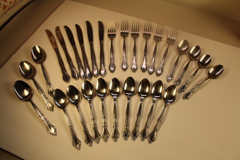 May include: A set of silverware with a silver finish, including forks, knives, and spoons. The silverware is arranged in a fan shape on a light brown surface.