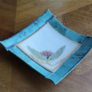May include: A square ceramic dish with a teal blue glaze and a raised rim. The dish features a hand-painted floral design in shades of yellow, green, and pink.