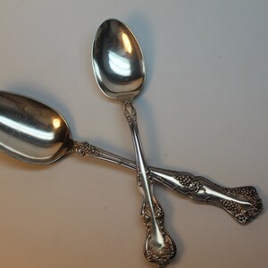 May include: Two silver spoons with ornate handles are crossed against a white background. The spoons have a classic design with a detailed floral pattern on the handles. The spoons are reflective, showing the light.