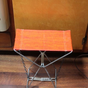 May include: A small, folding stool with an orange fabric seat and a silver metal frame. The seat is rectangular and has white stitching along the edges. The legs are connected by chains. The stool is approximately 30 cm tall.
