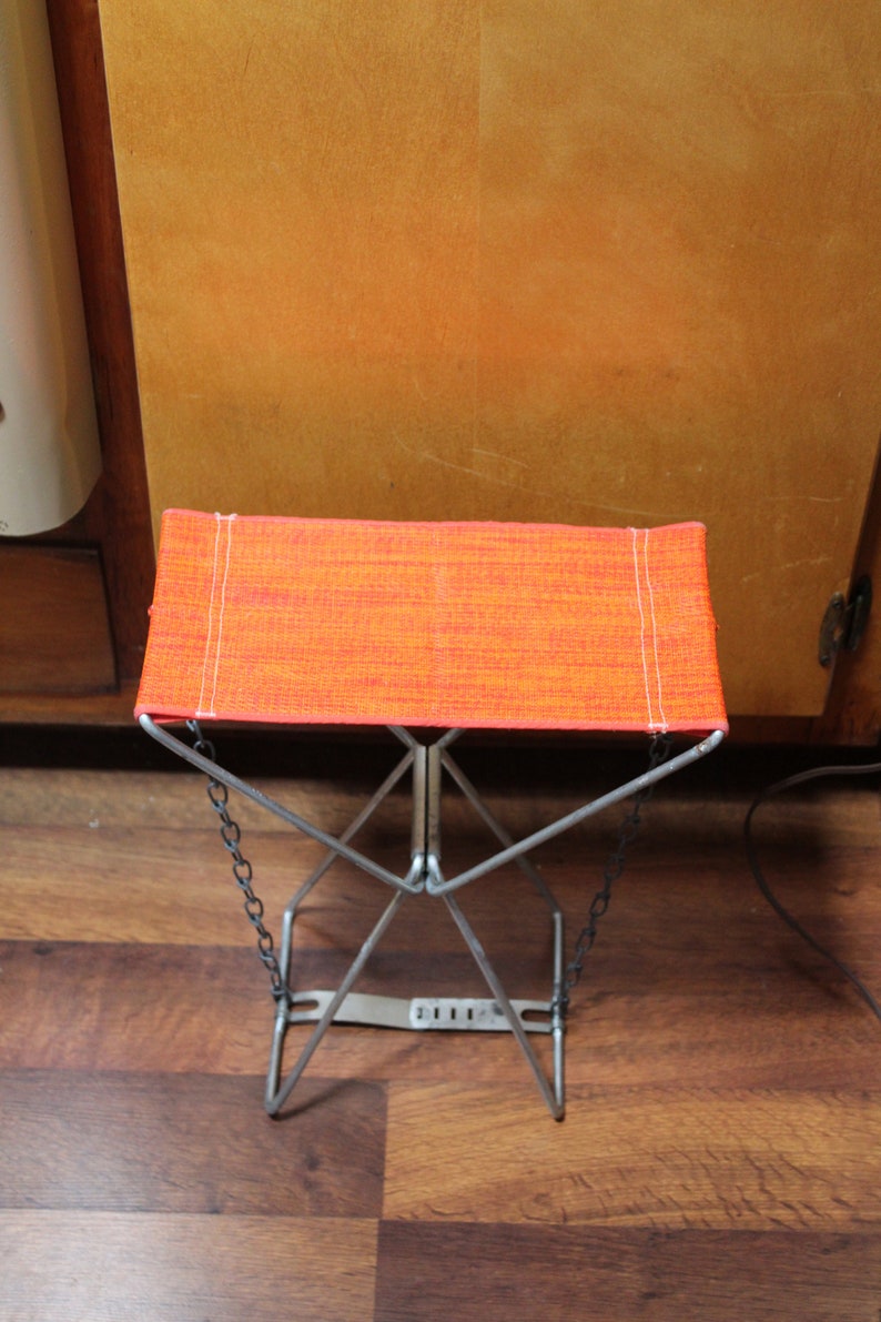 May include: A small, folding stool with an orange fabric seat and a silver metal frame. The seat is rectangular and has white stitching along the edges. The legs are connected by chains. The stool is approximately 30 cm tall.