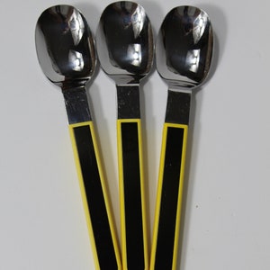May include: Three stainless steel spoons with a reflective surface. The handles are black with a yellow border. The spoons are arranged on a white surface.