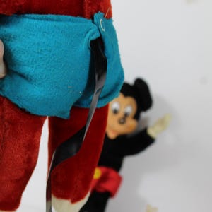 May include: A vintage Mickey Mouse plush toy, featuring red pants, a blue shorts, and a black tail. The toy has a black head and ears, with a smiling face. Other Mickey Mouse toys are in the background.