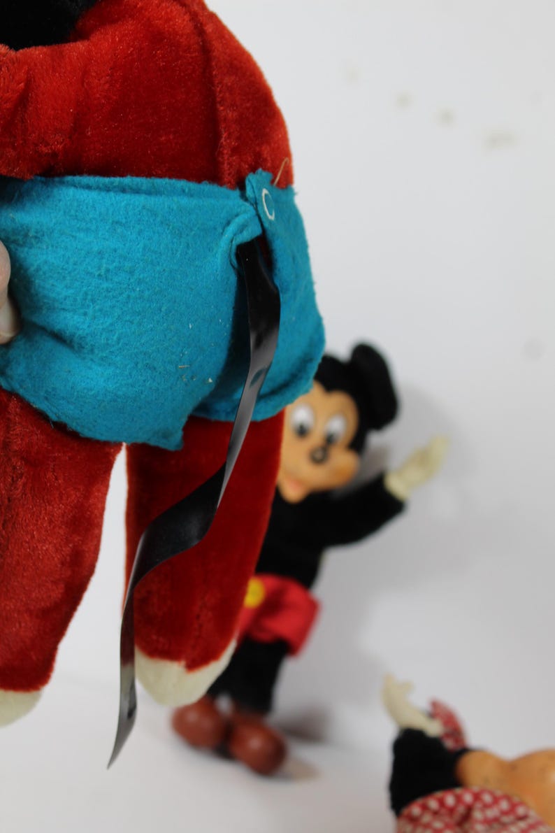 May include: A vintage Mickey Mouse plush toy, featuring red pants, a blue shorts, and a black tail. The toy has a black head and ears, with a smiling face. Other Mickey Mouse toys are in the background.