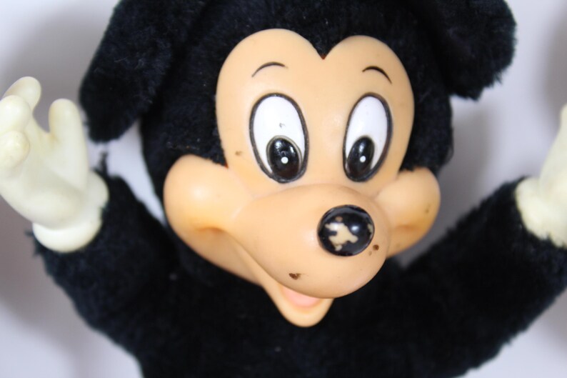 May include: Vintage Mickey Mouse plush toy with black fur, a tan face, and white gloves. The toy has large black eyes and a black nose. The plush is in a classic pose with arms outstretched.