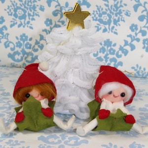 May include: A festive Christmas scene featuring two elf ornaments with red hats and green outfits, alongside a white, fluffy Christmas tree topped with a gold star. The ornaments have smiling faces and are seated. The background has a blue floral pattern.