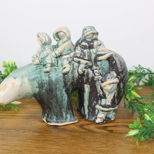 May include: A ceramic sculpture of a bear with three people riding on its back. The bear is green, brown, and white. The people are wearing winter clothing and are brown, white, and green.