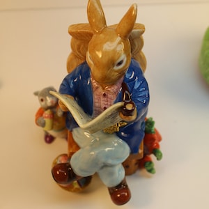 May include: A ceramic figurine of a brown rabbit wearing a blue jacket, reading a newspaper and holding a pipe. The rabbit is sitting on a chair with a smaller rabbit figurine standing next to it.
