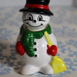 May include: A ceramic snowman figurine with a black hat, red scarf, and red mittens. The snowman has a yellow broom and black button details. A festive holiday decoration.