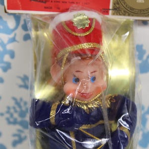 May include: A vintage Christmas ornament of a toy soldier in a plastic package. The soldier wears a red hat with gold trim, a blue uniform with gold accents, and has a smiling face with blue eyes. The packaging has a red and gold label.