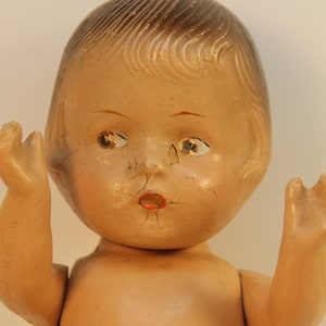 May include: A vintage baby doll with a painted face and arms raised. The doll has a light skin tone, with cracked paint on the face and a red painted mouth. The doll's hair is painted a light brown color.