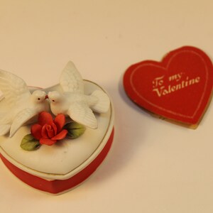 May include: A heart-shaped trinket box with a red base and a white lid, adorned with two white doves and a red rose. A red heart-shaped card with "To my Valentine" text is next to the box. The items are likely Valentine's Day themed.