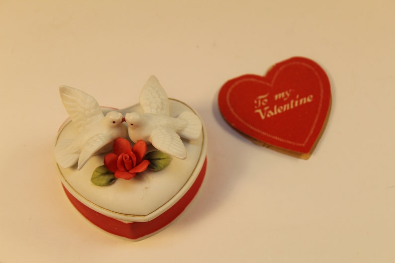 May include: A heart-shaped trinket box with a red base and a white lid, adorned with two white doves and a red rose. A red heart-shaped card with "To my Valentine" text is next to the box. The items are likely Valentine's Day themed.