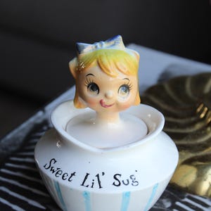May include: A vintage ceramic sugar bowl with a smiling girl figurine lid. The bowl is white with blue vertical stripes and the words "Sweet Lil' Sug" in black script. The girl has blonde hair, a blue bow, and is sticking out her tongue.