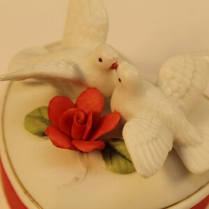 May include: A heart-shaped trinket box with a red base and a white lid. Two white doves are perched on top, with a red rose and green leaves. The box has a gold trim.