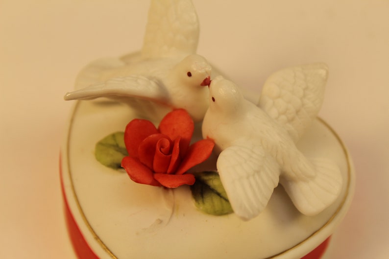 May include: A heart-shaped trinket box with a red base and a white lid. Two white doves are perched on top, with a red rose and green leaves. The box has a gold trim.