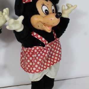 May include: A vintage Minnie Mouse plush doll. The doll has a black plush body, a red and white polka dot bow and skirt, and yellow shoes. The face is made of a tan plastic material with black eyes and a black nose.