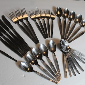 May include: A collection of stainless steel silverware, including forks, knives, and spoons, arranged on a white surface. The silverware has a simple, elegant design, with long, slender handles and rounded spoon bowls. The arrangement suggests a formal dining setting.