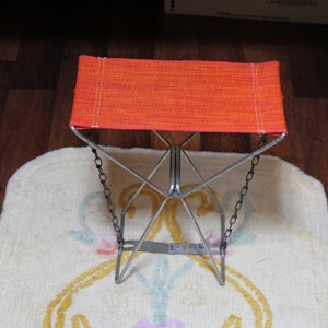 May include: An orange folding stool with a metal frame and chain accents. The seat is made of a woven orange fabric. The stool is on a cream-coloured rug with a floral design.