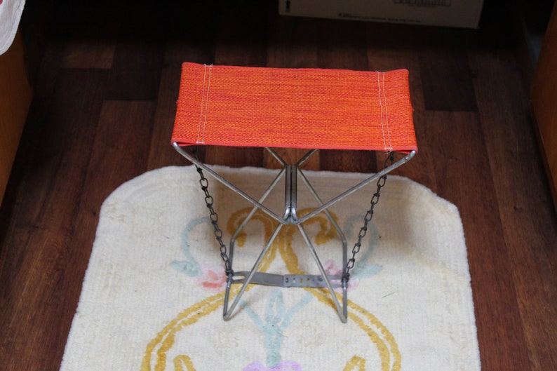 May include: An orange folding stool with a metal frame and chain accents. The seat is made of a woven orange fabric. The stool is on a cream-coloured rug with a floral design.
