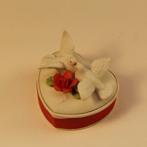 May include: A heart-shaped trinket box with a red base and a white lid. Two white doves are perched on top, with a red rose and green leaves. The box has a gold trim.