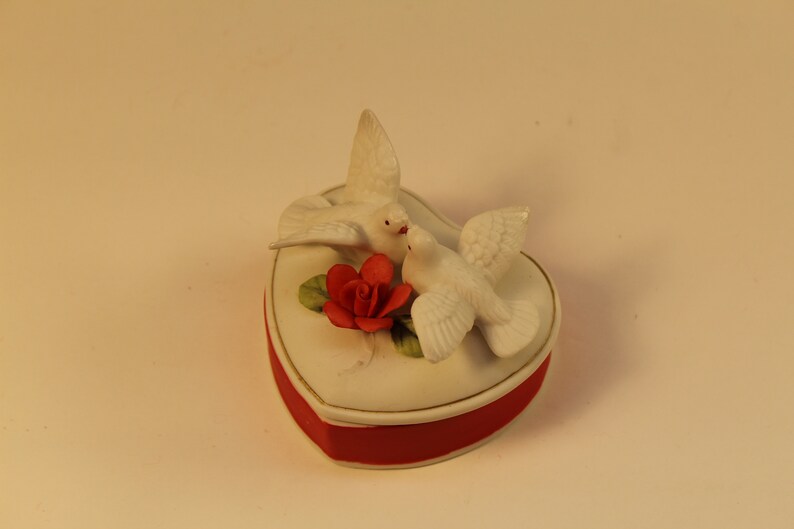 May include: A heart-shaped trinket box with a red base and a white lid. Two white doves are perched on top, with a red rose and green leaves. The box has a gold trim.