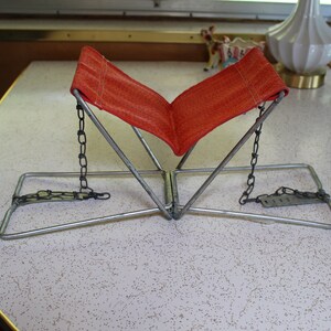 May include: An orange fabric book stand with a metal frame and chain supports. The stand is open, ready to hold a book. The stand is on a speckled white table.