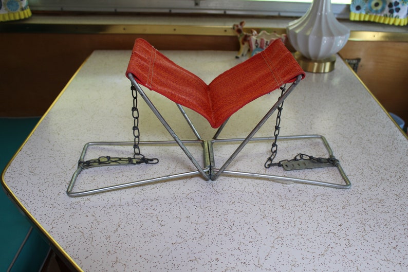 May include: An orange fabric book stand with a metal frame and chain supports. The stand is open, ready to hold a book. The stand is on a speckled white table.