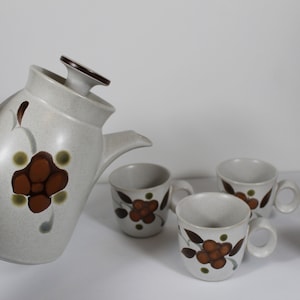 May include: A white ceramic teapot with a brown floral design is pouring tea into a set of four white ceramic teacups with brown floral designs.